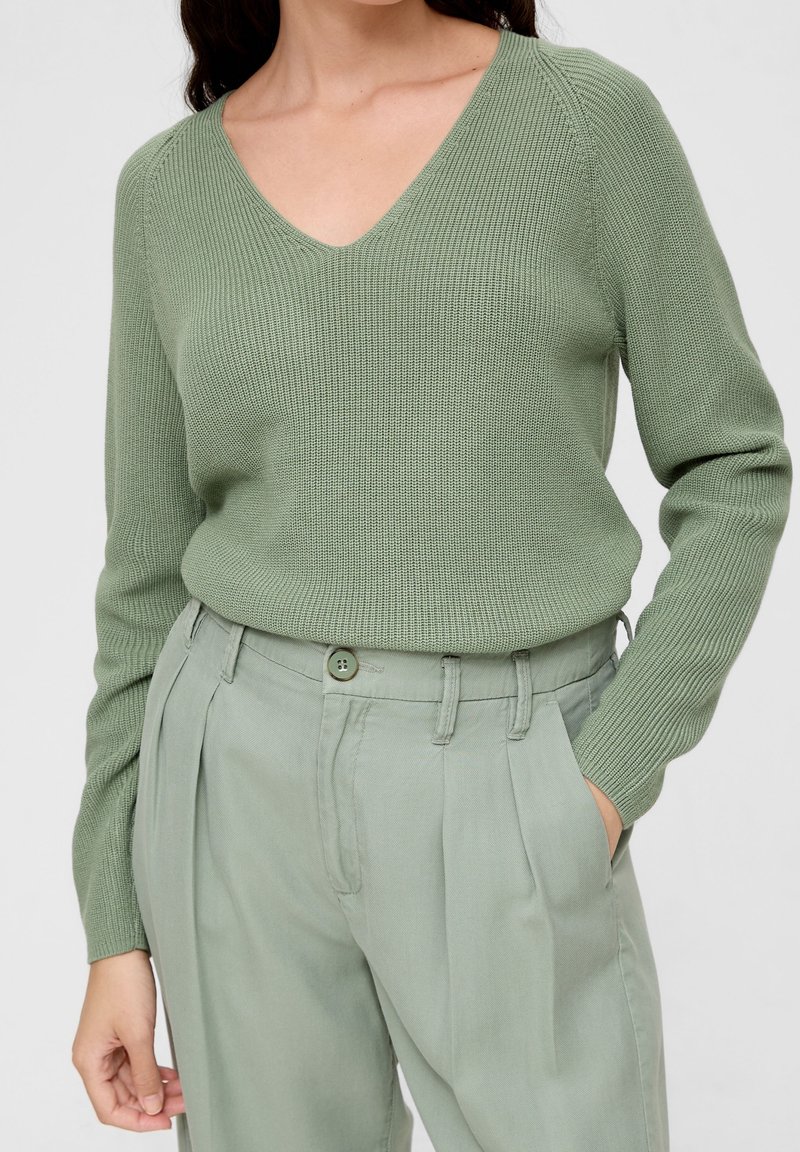 Maglione verde a V con una texture a coste, abbinato a pantaloni plissettati verde chiaro. Il maglione ha maniche lunghe e vita aderente.