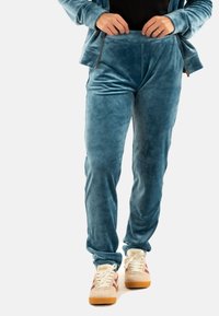 Personne portant un pantalon de jogging en velours bleu, des baskets beiges avec des rayures maroon, et une veste zippée bleue assortie, ajustant la taille.