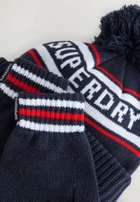 Námořnická modrá pletená čepice s pomponem a odpovídající rukavice, obě s bílými a červenými pruhy a štítky s logem "Superdry".