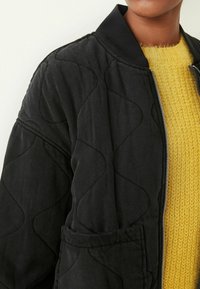 Czarna pikowana kurtka bomberka z prążkowanym kołnierzem i boczną kieszenią, nałożona na teksturowany żółty sweter z dzianiny. Widoczne detale srebrnego zamka.