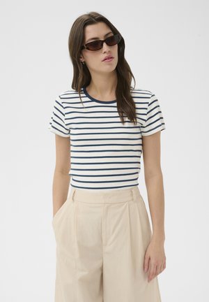 Femme portant des lunettes de soleil foncées, un t-shirt à manches courtes rayé blanc et bleu marine, et un pantalon beige taille haute, les mains dans les poches.