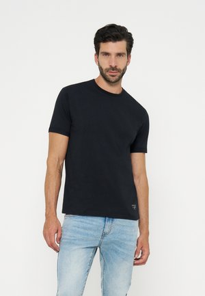 Man with dark hair and beard wearing a plain black short-sleeve t-shirt and light blue jeans, standing against a white background.