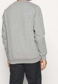 Hombre vestido con una sudadera de manga larga gris clara y pantalones negros, visto desde atrás contra un fondo blanco.