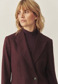 Burgundský blejzr s klopovým límcem, zapínání na jeden knoflík a jemnou texturou. Nosený přes tmavý rolák.