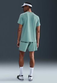 Mannelijke atleet in lichtgroene sportkleding, witte pet en witte sokken met zwart-witte sneakers, staande op een grijze studiovloer met zijn rug naar de camera gekeerd.
