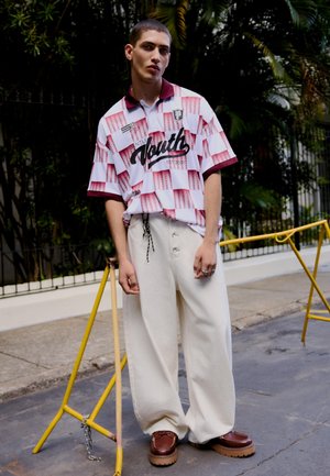 Mladý muž stojaci na ulici, mierne sa opierajúci, oblečený v červeno-bielej kockovanej polo košeli, širokých béžových nohaviciach a hnedých topánkach.