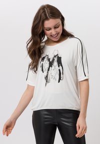 Weißes Kurzarmoberteil mit einem schwarz-weißen Grafikdruck von zwei Figuren, mit schwarzen Akzenten an den Schultern und einer weichen Stoffstruktur.