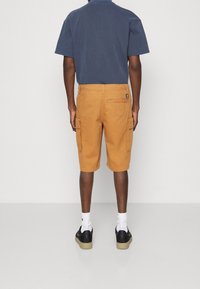 Marineblaues Kurzarmhemd kombiniert mit senfgelben Cargo-Shorts mit Seitentaschen. Schwarze Sneaker mit weißen Socken runden das Outfit ab.