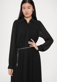 Γυναίκα ντυμένη με ένα μαύρο πλισέ φόρεμα με μακριά μανίκια και ασημένια ζώνη, στέκεται με το ένα χέρι στη μέση της μπροστά σε έναν απλό ανοιχτό φόντο.