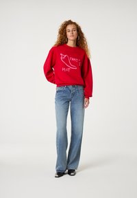 Rotes Sweatshirt mit pinkem Text und einer Chili-Pfeffer-Grafik, kombiniert mit hellblauen Schlagjeans und schwarzen Schuhen mit weißen Akzenten.
