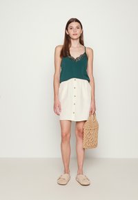 Camisola verde com rendas, combinada com uma saia creme de botões, segurando uma bolsa de palha, usando sapatos bégeis sem fecho.