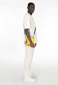 Gelbe Crossbody-Tasche mit schwarzem Reißverschluss, rechteckiger Form und erhabenem Schriftzug, getragen über einem weißen Outfit mit weißen Schuhen.