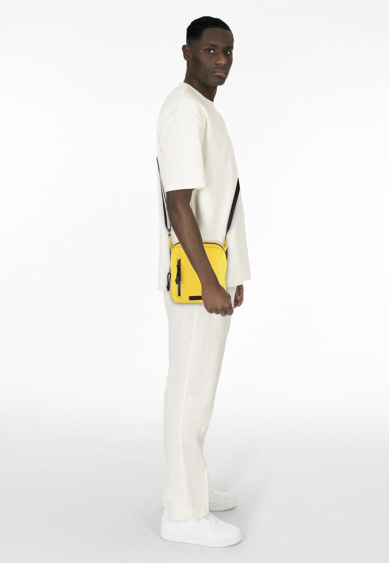 Gelbe Crossbody-Tasche mit schwarzem Reißverschluss, rechteckiger Form und erhabenem Schriftzug, getragen über einem weißen Outfit mit weißen Schuhen.