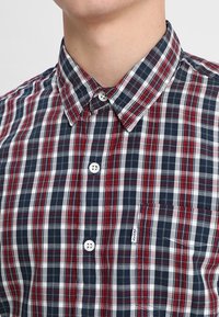 Chemise à carreaux en marine, rouge et blanc ; matière en coton ; col boutonné ; poche poitrine ; boutons blancs.