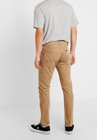 Slim-fit, béžové chinos vyrobené z odolné bavlny. Mají dvě zadní kapsy a středně vysoký pas. Kombinují se se šedým tričkem a černými teniskami.