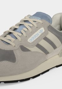 Ανοιχτό γκρι αθλητικό παπούτσι Adidas με κρεμ κορδόνια, μπεζ και γκρι ρίγες, και ανοιχτό μπλε λογότυπο στο πλάι και στη γλώσσα.