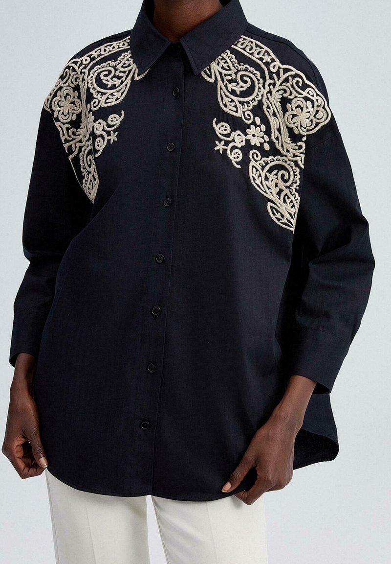 Chemise noire à manches longues boutonnée avec une broderie florale beige complexe sur les épaules, portée avec un pantalon blanc.