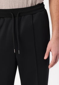 Schwarze Jogginghosen aus glattem Stoff mit Tunnelzug an der Taille. Verfügen über silberne Kordelenden und dezente Seitentaschen.