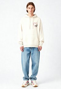 Felpa crema con un design a forma di cuore rosso, tasca frontale, polsini a coste, abbinata a jeans baggy azzurri e sneakers bianche con dettagli rossi.