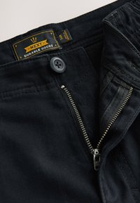 Pantaloni in denim nero con chiusura a cerniera e bottone. L'etichetta recita "NEXT DURABLE GOODS" in giallo. Presenta dettagli di cuciture fini.