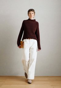 Maglione ribbed bordeaux, pantaloni bianchi a gamba larga e stivali alla caviglia beige. La modella tiene una pochette marrone. Sfondo semplice e pavimento in legno.