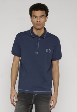 Jonge man staand, gekleed in een donkerblauw poloshirt met witte stiksels en een donkere spijkerbroek tegen een effen grijze achtergrond.