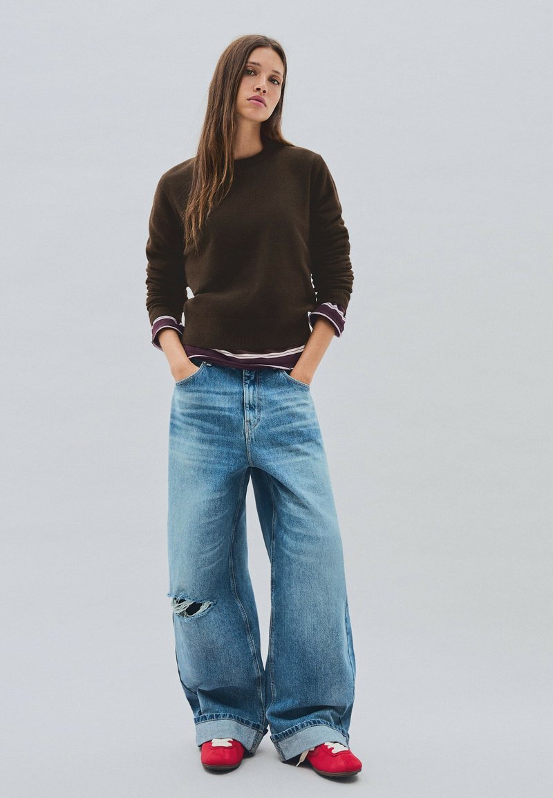 Jeune femme debout, mains dans les poches, portant un pull marron, une chemise superposée, un jean déchiré large et des baskets rouges sur un fond uni.