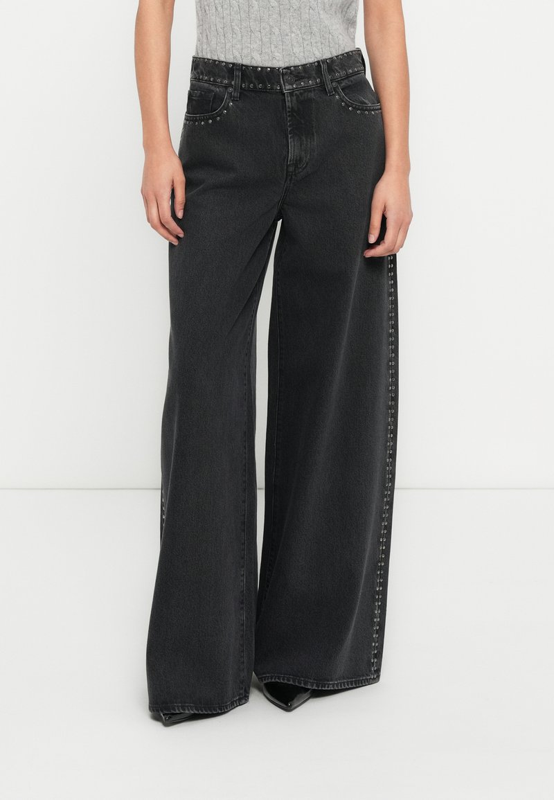 Μαύρο τζιν με φαρδιά πόδια και ψηλή μέση, με διακοσμητικά μεταλλικά καρφιά κατά μήκος των εμπρόσθιων τσέπων και των πλαϊνών ραφών, κατασκευασμένο από τζιν ύφασμα.