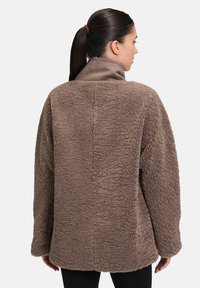 Brązowa, teksturowana polarowa kurtka z wysokim kołnierzem, w oversize'owym kroju i miękkim materiale, z długimi rękawami i bez widocznych zapięć.