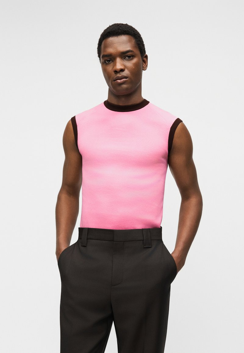 Man wearing sleeveless pink top with black trim and black pants, standing with hands in pockets against plain light background.