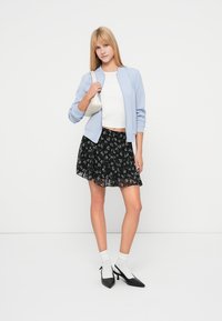 Veste bomber bleu clair portée par-dessus un haut blanc, associée à une jupe noire à motif floral. Portée avec des chaussettes blanches et des talons noirs.