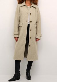 Beżowy płaszcz podszyty polarem, z kołnierzem, czterema przednimi kieszeniami i zapięciem na guziki. Długie rękawy z podwiniętymi mankietami, zestawione z czarnymi dżinsami i botkami na kostkę.