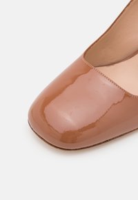 Pompe en cuir verni marron avec un bout carré et une texture lisse. Présente un devant échancré et une couture discrète le long du bord.