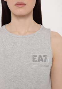 Szary bezrękawnik z okrągłym dekoltem i błyszczącą naszywką z logo "EA7" w srebrnym brokacie oraz napisem "EMPORIO ARMANI" poniżej.