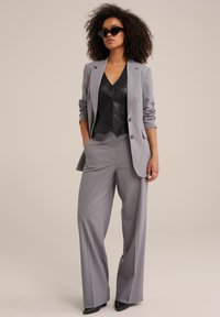 Traje gris a medida con pantalones de pierna ancha, blazer de un botón y camisón de satén negro. La modelo lleva gafas de sol negras, mostrando un diseño elegante.