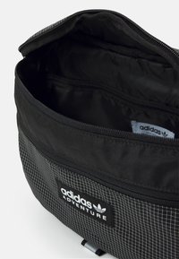 Svart Adidas Adventure-väska med vitt rutmönster, öppen toppdragkedja som visar det tomma inuti och Adidas-logotypen inuti.