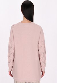 Helles rosa gestricktes Sweatshirt mit lockerem Schnitt. Merkmale sind rautenförmige Ausschnittmuster an den Ärmeln sowie ein gerippter Hals- und Armabschluss.