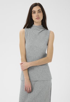 Femme aux longs cheveux foncés portant un haut gris sans manches côtelé et une jupe assortie, debout avec un bras croisé et une expression neutre.