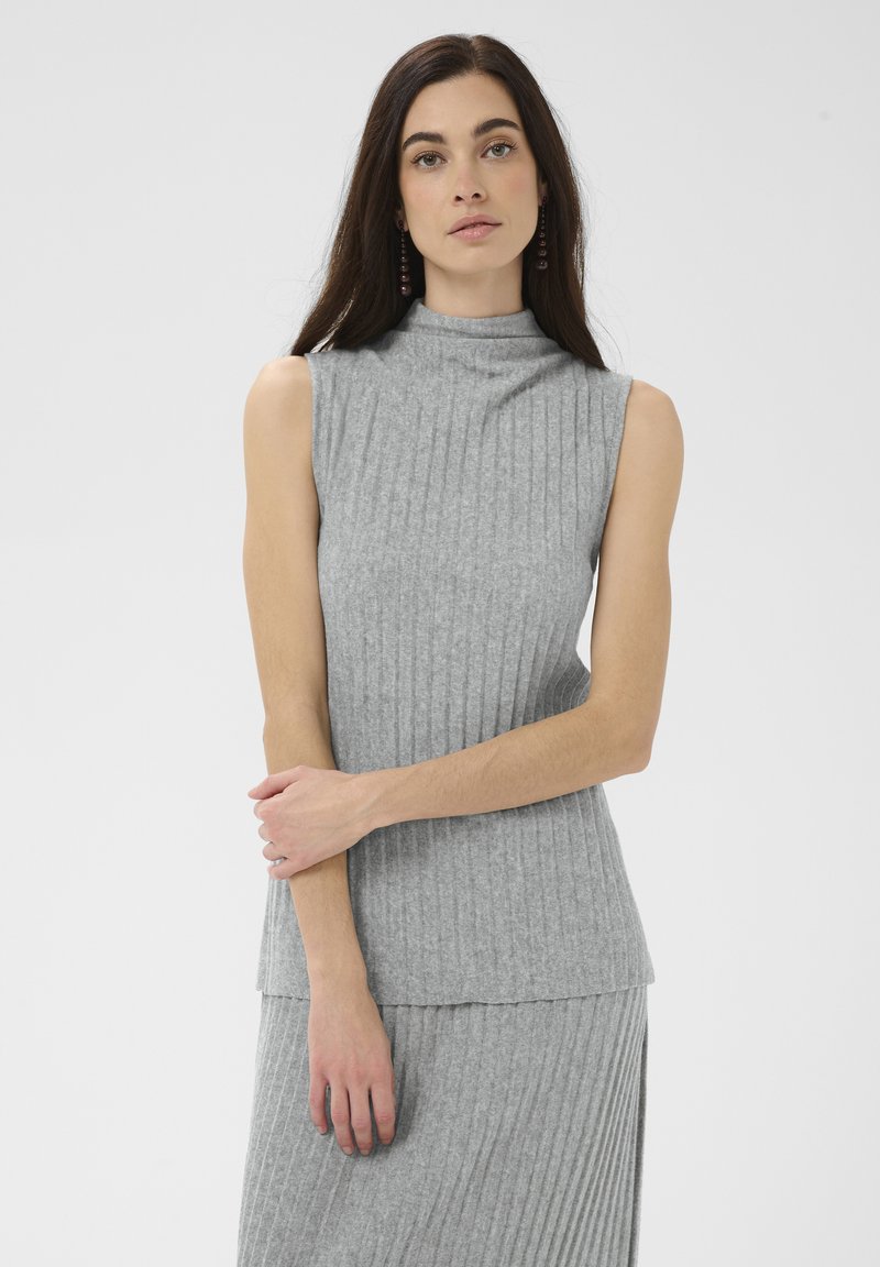 Femme aux longs cheveux foncés portant un haut gris sans manches côtelé et une jupe assortie, debout avec un bras croisé et une expression neutre.