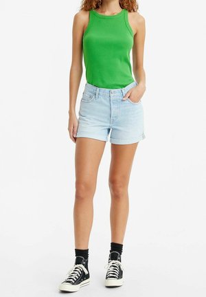 Femme portant un haut vert sans manches, un short en jean bleu clair roulé, des chaussettes noires et des baskets montantes noires, debout avec une main dans la poche.