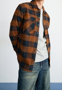 Camisa de franela a cuadros marrón y negro con dos bolsillos delanteros, cierre de botones y cuello con solapa, combinada sobre una camiseta gris.