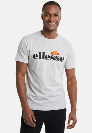 Homme souriant portant un t-shirt ellesse gris clair avec un pantalon noir, debout devant un fond blanc uni.