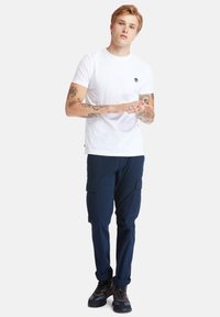 Weißes Baumwoll-T-Shirt mit kleinem Logo, kombiniert mit marineblauen Cargo-Hosen mit mehreren Taschen und robusten, dunklen Schuhen.