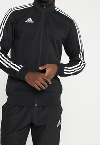 Svart Adidas jacka med dragkedja och hög krage, med tre vita ränder längs varje ärm och en liten vit logotyp på bröstet.