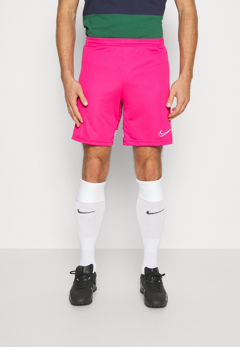 Homme portant un short de sport Nike rose vif, des chaussettes montantes blanches Nike et des crampons noirs, debout devant un fond blanc uni.