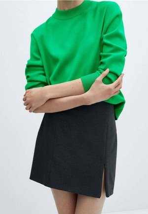 Persona vistiendo un suéter verde brillante y una falda gris oscuro con una pequeña abertura lateral, con los brazos cruzados suavemente a la altura de la cintura.