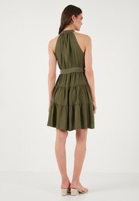 Abito verde oliva senza maniche con gonna a strati, vita cinte e tessuto morbido. Presenta un collo plissettato e una vestibilità rilassata.