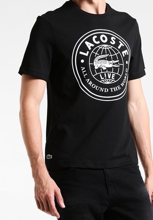 Czarny krótki rękaw T-shirt wykonany z bawełny, z dużym białym okrągłym logo przedstawiającym grafikę krokodyla oraz tekstem poniżej.