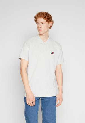 Polo de color gris claro con cuello, mangas cortas y un pequeño logo bordado. La tela parece suave, con un corte holgado.