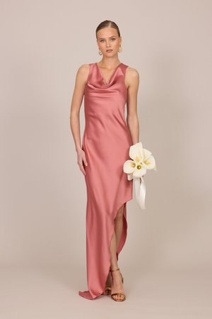 Frau in einem ärmellosen rosa Satinkleid mit asymmetrischem Saum, die einen weißen Calla-Lilien-Strauß hält und goldene Absätze sowie Creolen trägt.